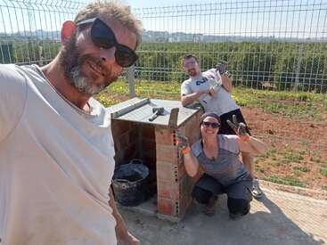 Three people pose cheerfully at a construction site, with one taking a selfie. They are building with bricks, wearing gloves, and enjoying the sunny outdoor environment.