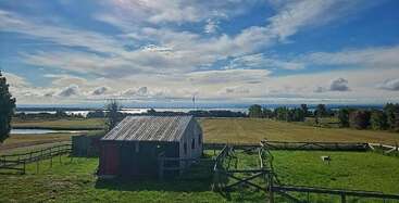The image depicts a serene rural landscape featuring a small farmhouse with a fenced yard, surrounded by rolling hills and a tranquil body of water, evoking a sense of peacefulness and tranquility.