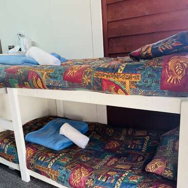 This image shows a set of white wooden bunk beds with colorful patterned bedding. Each bed has pillows, a blue towel, and a rolled white towel.