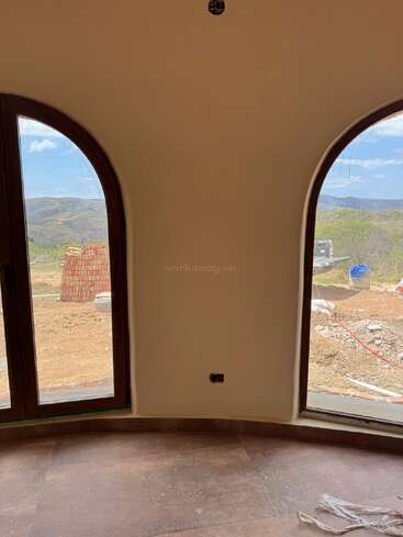 Dieses Bild zeigt zwei gewölbte Fenster in einem beigefarbenen Raum mit Blick auf eine malerische Landschaft mit Hügeln, Baumaterialien, einem Auto und blauem Himmel.