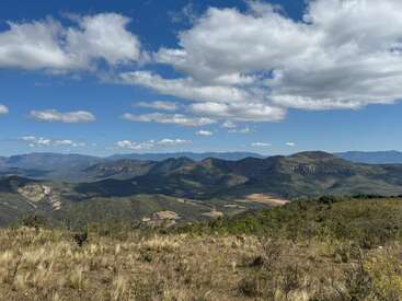 Sanfte Berge erstrecken sich unter einem strahlend blauen Himmel, der mit weißen Wolken übersät ist. Trockenes Gras und Sträucher bedecken den Vordergrund und schaffen eine friedliche, weitläufige Landschaft.