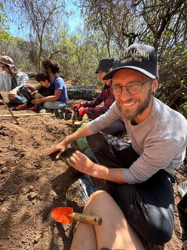 Eine Gruppe von Menschen, darunter ein lächelnder Mann mit Brille und Mütze, ist im Freien, umgeben von Bäumen und Sonnenlicht, mit Garten- oder Pflanzarbeiten beschäftigt.