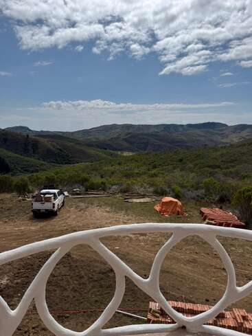Ein weißes Geländer blickt auf eine ländliche Szene mit einem Pickup, gestapelten Ziegeln und einer Plane. Grüne Hügel und Berge erstrecken sich unter einem teilweise bewölkten Himmel.