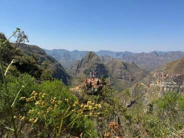 Eine atemberaubende Berglandschaft mit üppigem Grün und schroffen Gipfeln unter einem klaren blauen Himmel. Zwei Menschen stehen auf einer Klippe und genießen den Panoramablick.
