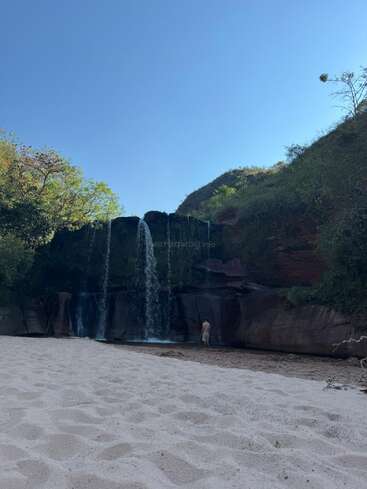 Eine ruhige Sandfläche führt zu einem stillen Wasserfall, der über dunkle Felsen stürzt, umgeben von üppigem Grün und einem klaren, strahlend blauen Himmel.