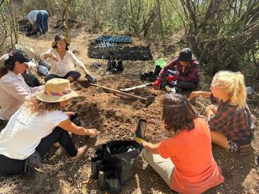 Un groupe de personnes est réuni en plein air, assis et agenouillé en cercle, travaillant avec de la terre et du matériel de plantation, entouré d'arbres et de bacs à semis.