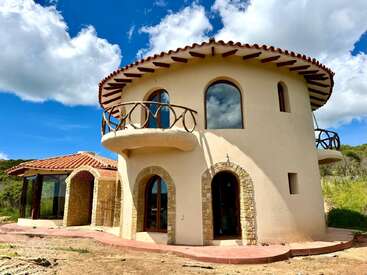 This is a charming, round, Mediterranean-style house with arched windows and doors, stone accents, balconies, and a terracotta-tiled roof under a bright sky.
