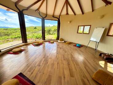 A bright, circular room with wooden floors, large windows overlooking nature, meditation cushions arranged in a circle, and a whiteboard, creating a peaceful, inviting atmosphere.