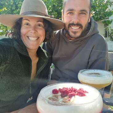 Ein lächelndes Paar sitzt draußen und genießt Cocktails in der Sonne. Die Frau hat einen Hut mit breiter Krempe und einen grünen Fleecepulli an und fängt einen fröhlichen, entspannten Moment zu zweit ein.