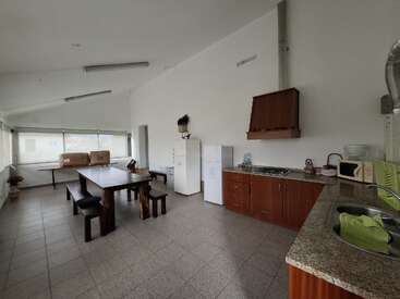 This image shows a spacious kitchen with tiled flooring, wooden cabinets, a long dining table, benches, refrigerator, microwave, sink, and large windows providing natural light.