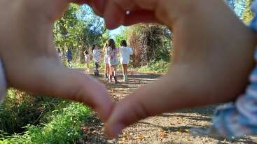Several children walk together down a sunlit forest path, framed by hands forming a heart shape in the foreground, symbolizing love, friendship, and adventure outdoors.