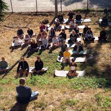 A group of people sits outdoors on mats, arranged in rows on dry grass, attentively facing an individual at the front, enjoying sunlight and nature.