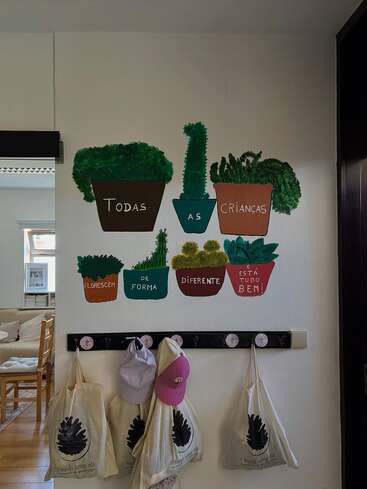 This image shows a wall decorated with painted potted plants and a Portuguese phrase about children. Four bags and two hats hang on hooks below. Cozy, inviting space.