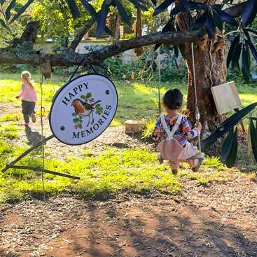 Zwei Kinder spielen im Freien in der Nähe eines Baumes. Das eine schaukelt fröhlich, während das andere rennt. Auf einem Schild am Ast steht "Happy Memories". Das Sonnenlicht fällt durch die Blätter.