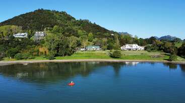 A peaceful lakeside scene with a small boat in the water, surrounded by lush green hills, trees, and charming houses under a clear blue sky.