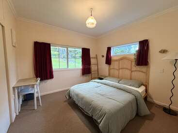 This cozy bedroom features a double bed, light green bedding, wooden headboard, two windows with red curtains, a small desk, chair, and standing lamp.