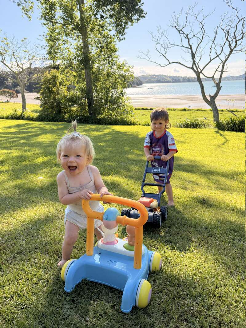 Two young children play joyfully on green grass near a beach. One pushes a colorful walker, the other uses a toy lawnmower. Trees and sunshine complete the scene.
