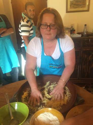 The image depicts a woman in an apron kneading dough on a wooden table, surrounded by bowls and utensils, with two people in the background wearing matching aprons.