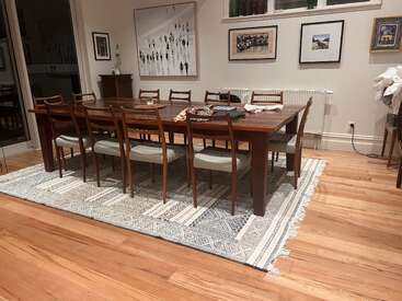 The image depicts a dining room with a wooden table and chairs on a rug, featuring artwork on the walls and a radiator against the wall.