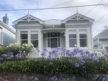 The image depicts a charming house with a white picket fence, surrounded by vibrant purple flowers and lush greenery, set against a serene blue sky.