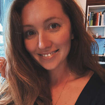 Eine Frau mit langen braunen Haaren lächelt in die Kamera. Sie ist drinnen, mit Bücherregalen und einem blauen Stuhl im Hintergrund. Das Licht ist warm.