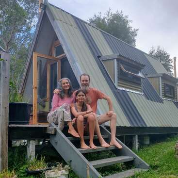 Uma família de três pessoas senta-se descalça nos degraus de uma pequena e moderna cabana com estrutura em A, cercada por vegetação, sorrindo satisfeita para a câmera.