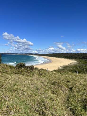 A pristine, sandy beach curves along the turquoise coastline, bordered by lush greenery. Fluffy clouds drift across a clear blue sky, creating a tranquil, scenic view.