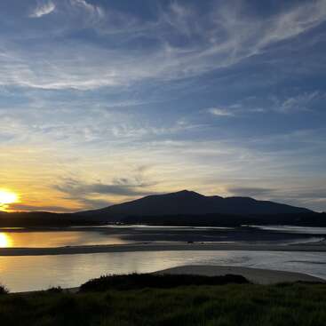 The image depicts a serene sunset over a body of water, with a mountain range in the background and a grassy bank in the foreground.