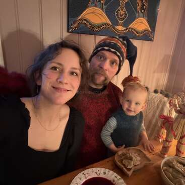 A family of three sits at a cozy wooden table, smiling warmly. Their meal and traditional decorations suggest a festive atmosphere in a warmly lit room.
