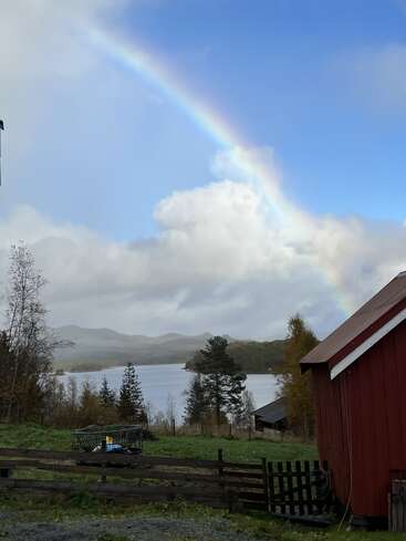 Ein leuchtender Regenbogen spannt sich über einen teilweise bewölkten Himmel über einem ruhigen See, umgeben von herbstlichen Bäumen, Bergen, einem rustikalen Zaun und einer roten Scheune.