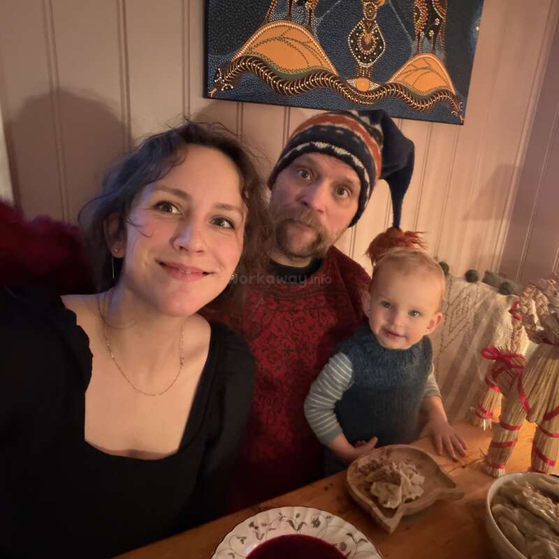 A family of three sits at a cozy wooden table, smiling warmly. Their meal and traditional decorations suggest a festive atmosphere in a warmly lit room.