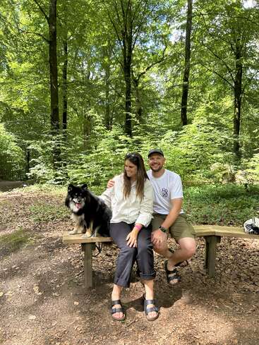 Un couple est assis sur un banc en bois dans une forêt luxuriante et ensoleillée. Ils sourient, profitant de l'extérieur avec leur chien noir heureux à leurs côtés. Scène paisible.