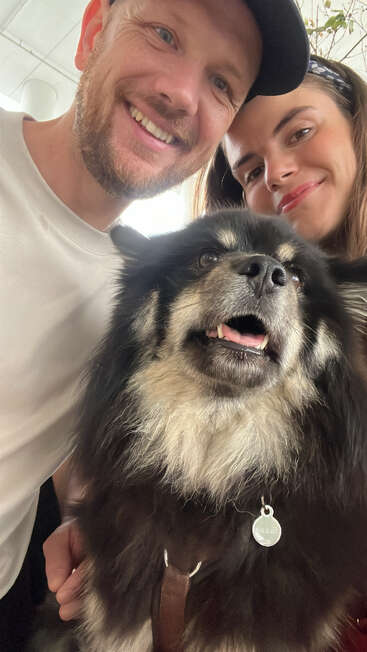 A joyful man and woman smile for a close-up selfie with their fluffy black and white dog, who looks happy wearing a tag on its collar.