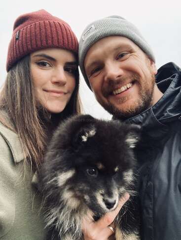 L'image montre un selfie d'un homme et d'une femme portant des bonnets d'hiver, avec un petit chien noir devant eux, sur fond de ciel gris.