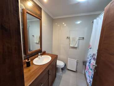 Este cuarto de baño cuenta con un tocador de madera con lavabo y espejo, inodoro, toallero, radiador y una ducha con cortina con motivos florales. Iluminación brillante.