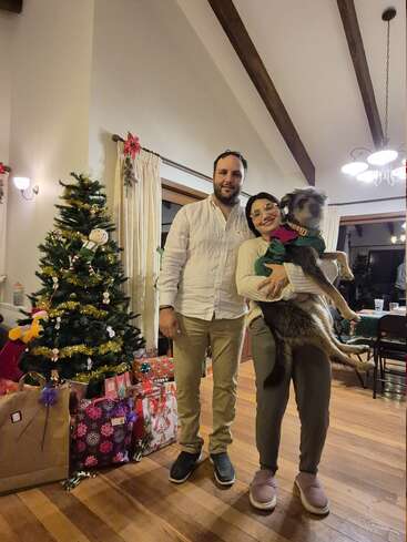 Una pareja posa en interiores junto a un árbol de Navidad decorado y muchos regalos. Una persona sostiene a un perro vestido con ropa festiva. Ambiente cálido y navideño con iluminación acogedora.