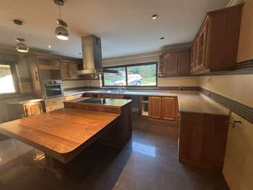 Esta espaciosa cocina cuenta con armarios de madera, una isla con fogones integrados, iluminación moderna, grandes ventanas con vistas al exterior y amplios espacios de encimera.