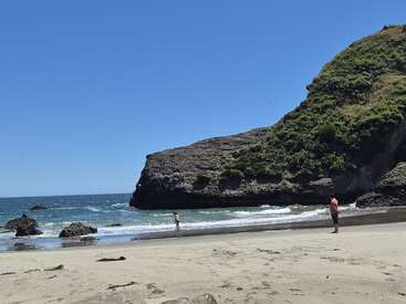 Una soleada escena playera muestra a dos personas cerca del agua, una costa rocosa, olas del mar y un gran acantilado rocoso cubierto de vegetación contra un cielo azul claro.