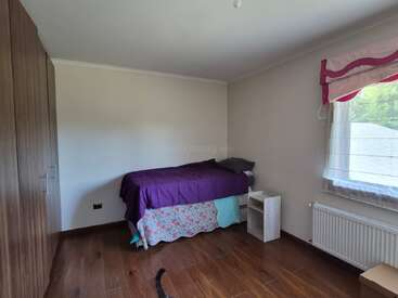 A simple bedroom with a single bed covered in purple bedding, a small wooden nightstand, wood flooring, plain walls, and a window with pink curtains.