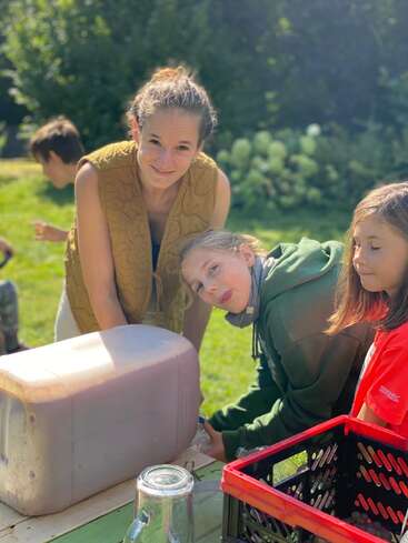 Three kids and an adult enjoy a sunny outdoor activity. They gather around a large water container, filling bottles, with smiles and curiosity visible on their faces.