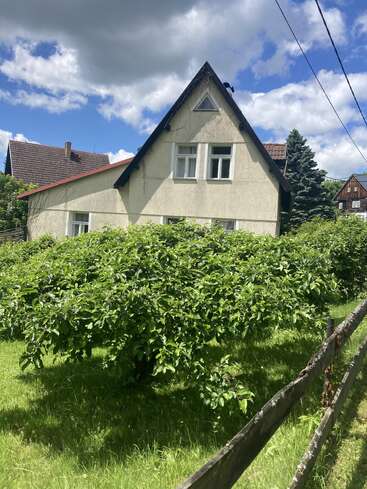 A quaint house with a triangular roof sits behind lush green trees and a wooden fence. The sky is partly cloudy, creating a peaceful atmosphere.