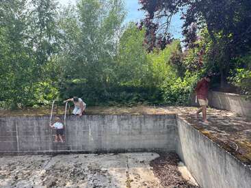 An empty, abandoned swimming pool with moss and leaves. A child climbs the ladder, assisted by an adult, while another person walks along the pool's edge. Lush greenery surrounds.