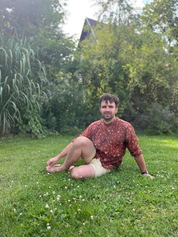 Un homme est assis pieds nus sur l'herbe verte, vêtu d'une chemise à fleurs rouge et d'un short beige. Entouré d'une végétation luxuriante, il profite d'un moment de tranquillité en plein air.