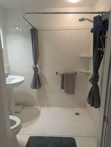 Das Bild zeigt ein sauberes, modernes Badezimmer mit einer begehbaren Dusche, grauen Duschvorhängen, einem Waschbecken mit Sockel, einer Toilette, einem Handtuchhalter, Regalen und einer blauen Badematte.