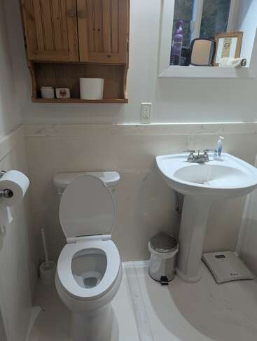 Auf dem Bild sieht man ein kleines, ordentliches Badezimmer mit Toilette, Waschbecken mit Unterschrank, Mülleimer, Waage, Holzschrank, Toilettenpapier und einem Fenster, auf dem verschiedene Sachen stehen.