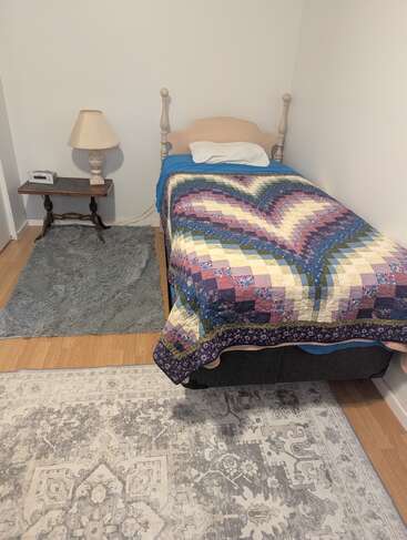 Das gemütliche Schlafzimmer hat ein Einzelbett mit einer bunten Bettdecke, ein Kissen, einen Nachttisch aus Holz mit einer Lampe, einen Wecker und zwei Teppiche.