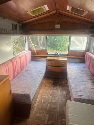 Cette image montre l'intérieur confortable d'un camping-car vintage, avec deux lits simples, des armoires en bois, de grandes fenêtres et un sol en linoléum à motifs.