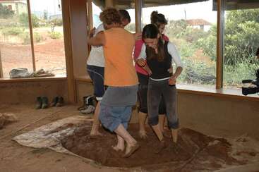 Quatro pessoas estão descalças, pisando e misturando lama dentro de casa sobre uma lona. Parecem estar se divertindo, talvez como parte de uma atividade ecológica ou em equipe.