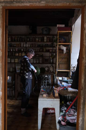 Una persona se encuentra en un taller acogedor y abarrotado, rodeada de estanterías llenas de tarros, herramientas y diversos suministros, concentrada en trabajar con el equipo.