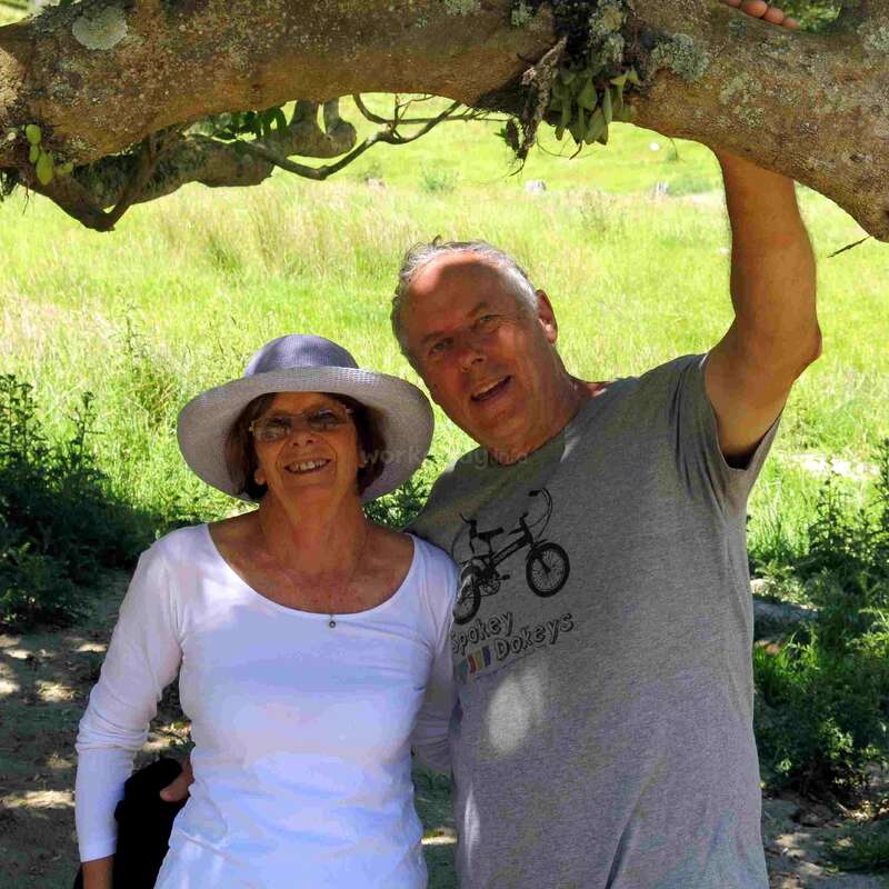 The image depicts an older couple standing together under a tree, with the woman wearing a white shirt and hat, and the man in a gray t-shirt, set against a grassy background.